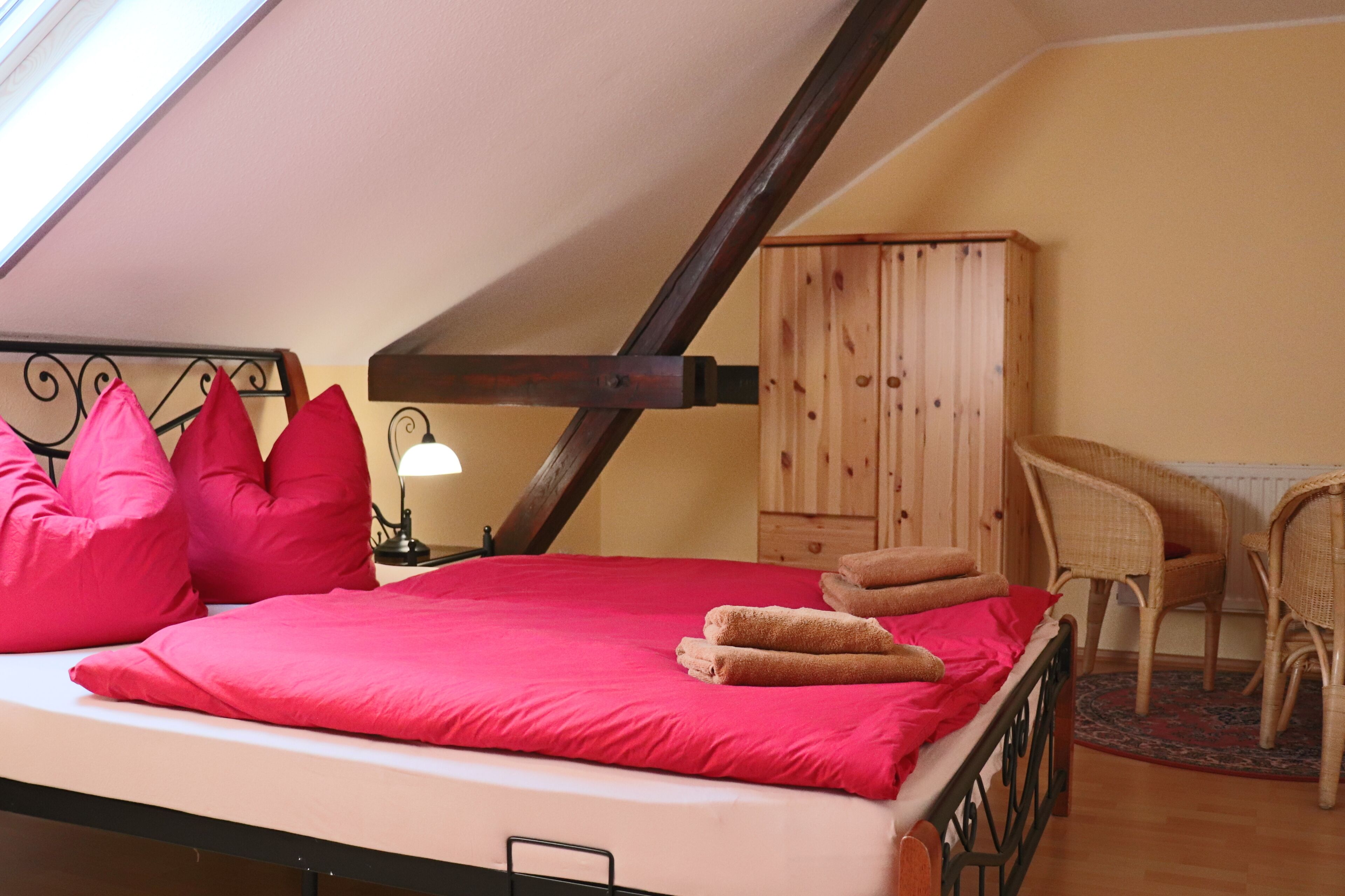 A bedroom with a sloped ceiling, a bed with red bedding, a wooden wardrobe, and a wicker chair.