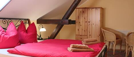 A bedroom with a sloped ceiling, a bed with red bedding, a wooden wardrobe, and a wicker chair.