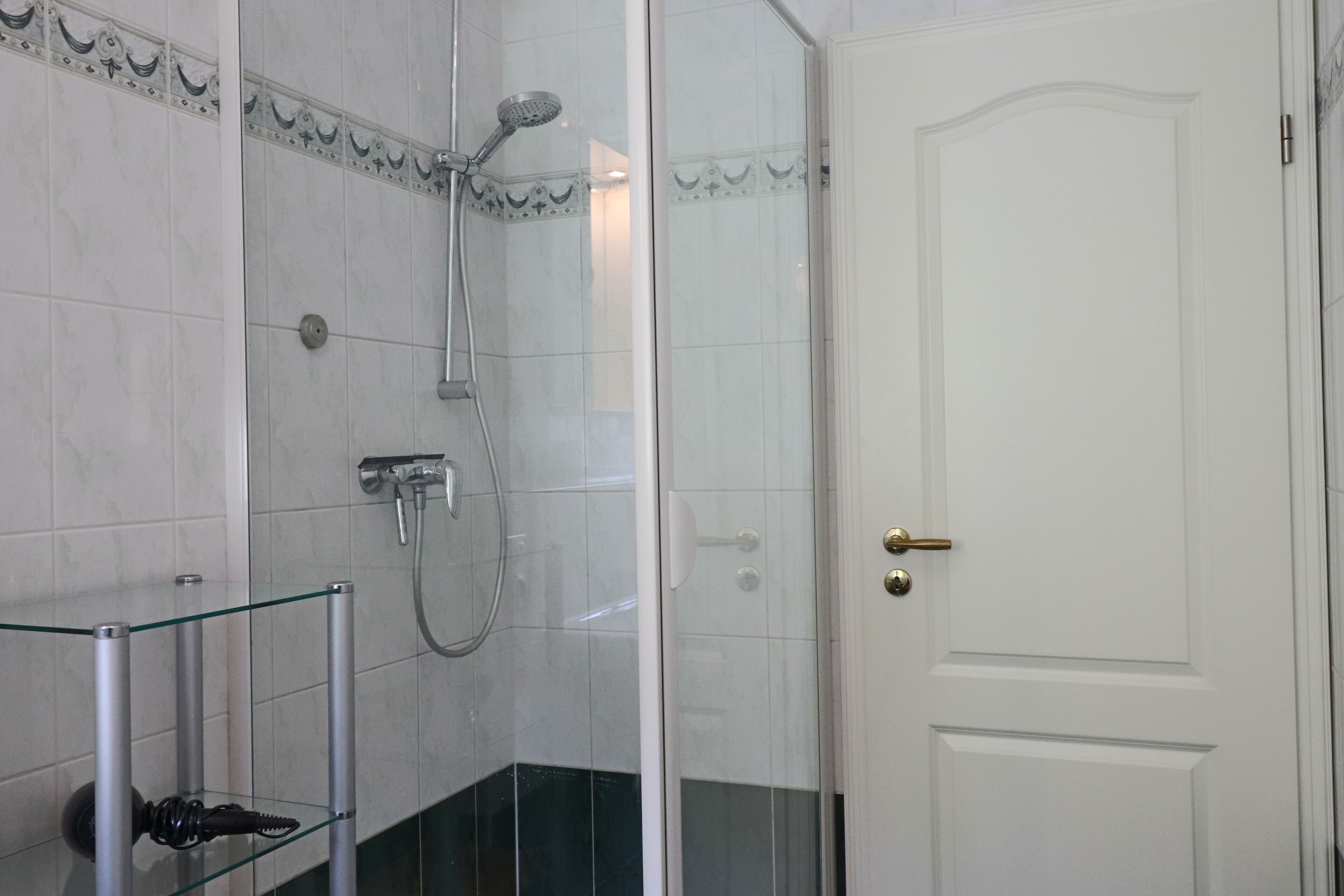 Une salle de bain avec une douche à parois de verre, une porte blanche avec une poignée en laiton et une étagère en verre sur laquelle repose un objet noir.