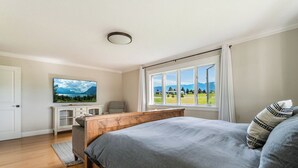 Un dormitorio con cama, televisor, ventana y vista a un paisaje verde, y cabecera de madera.