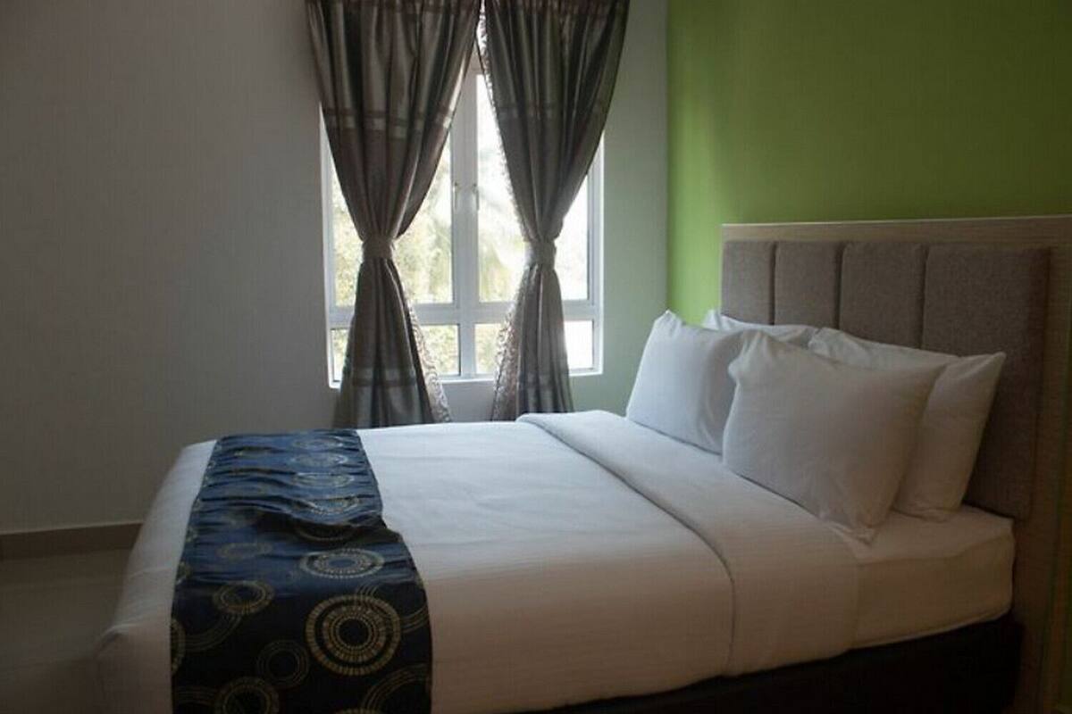 A neatly made bed with a patterned throw, a window with curtains, and a green wall.