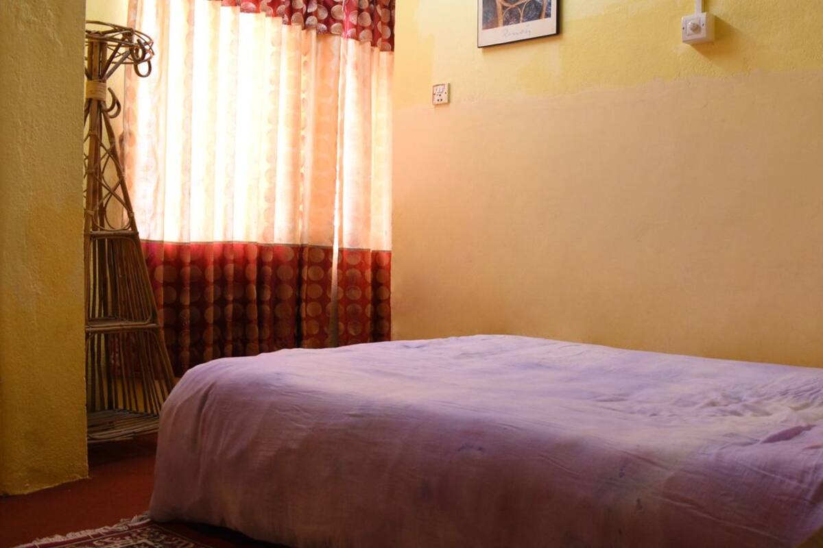A bedroom with a bed, a window with curtains, a wall-mounted light switch, and a wall-mounted picture.
