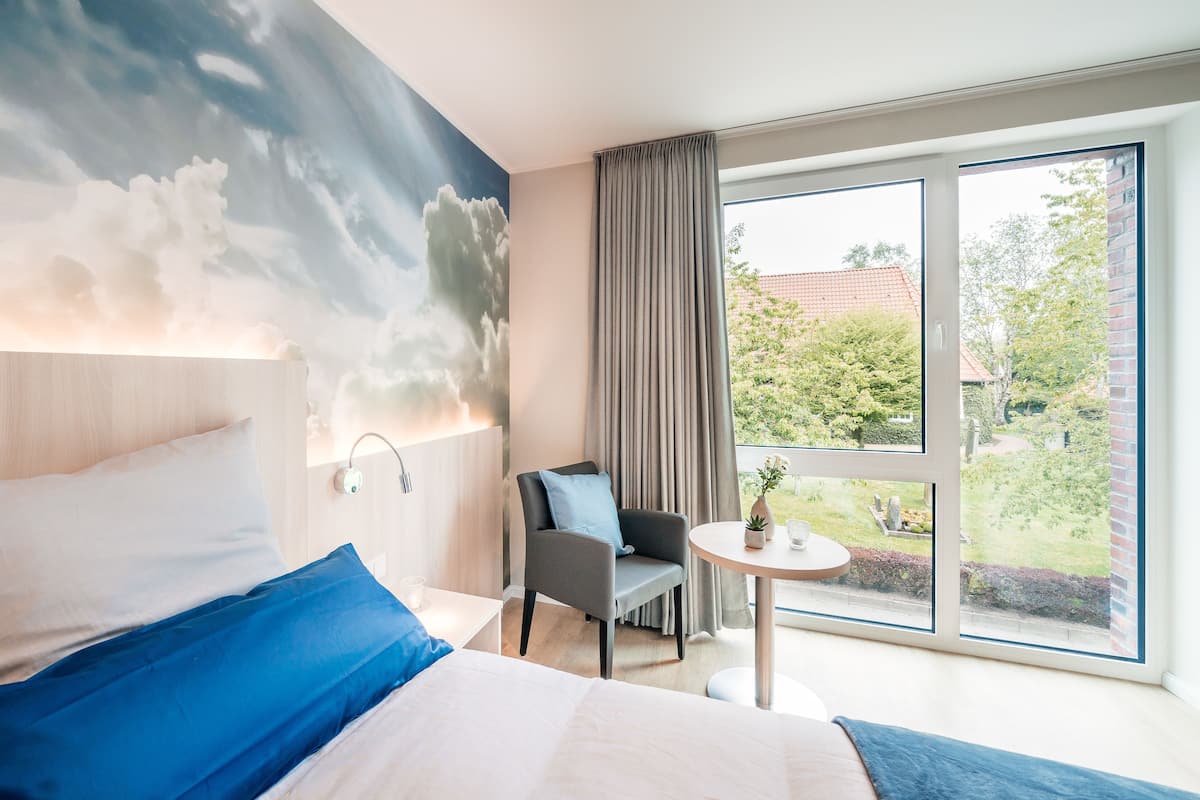 A modern hotel room with a large bed, a chair, a small table, and a view of the garden through a large window.