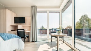 A modern hotel room with a bed, desk, chair, and a balcony with a table and plants.