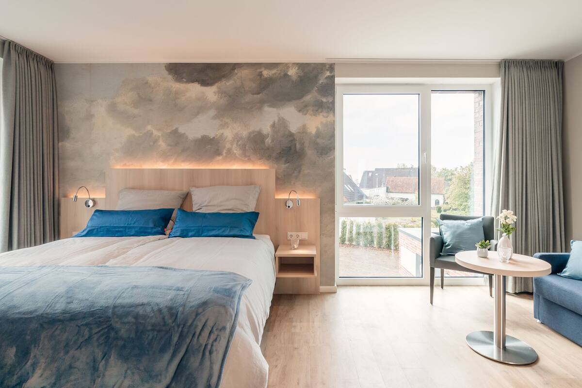 A modern hotel room with a large bed, a small table, a sofa, and a balcony with a view.