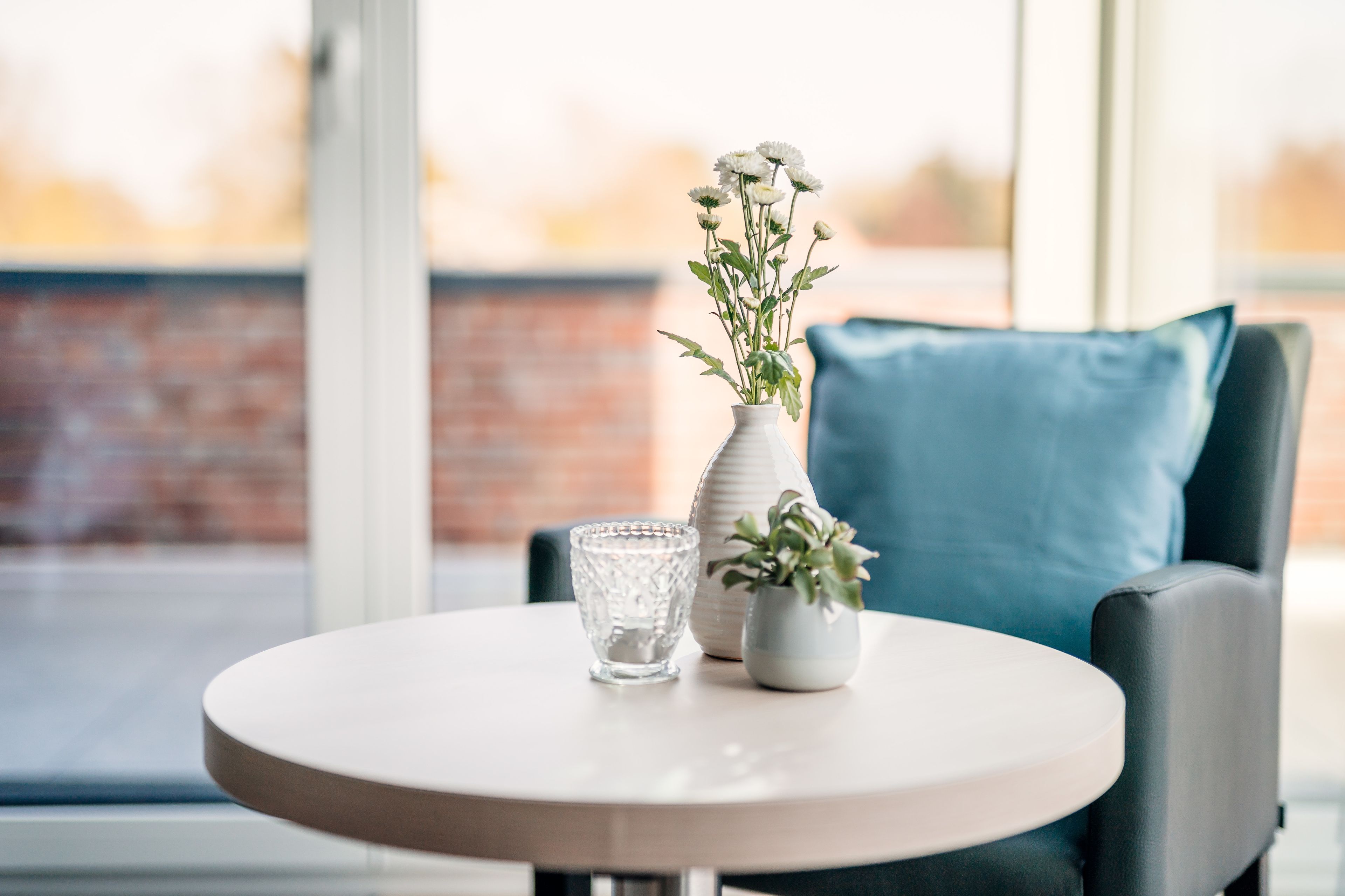 A round table with a vase of flowers and a glass on it, placed in front of a window with a brick wall in the background.