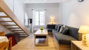 Een moderne woonkamer met een bank, een salontafel en een lamp. Links is er een trap en een raam met gordijnen.