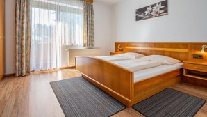 Ett hotellrum med en säng i trä, ett nattduksbord, en vägglampa och ett fönster med tunna, genomskinliga gardiner.