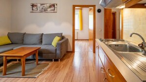 Ett kompakt bostadsutrymme med ett litet kök, en soffa med kuddar, ett soffbord i trä och en tavla på väggen.