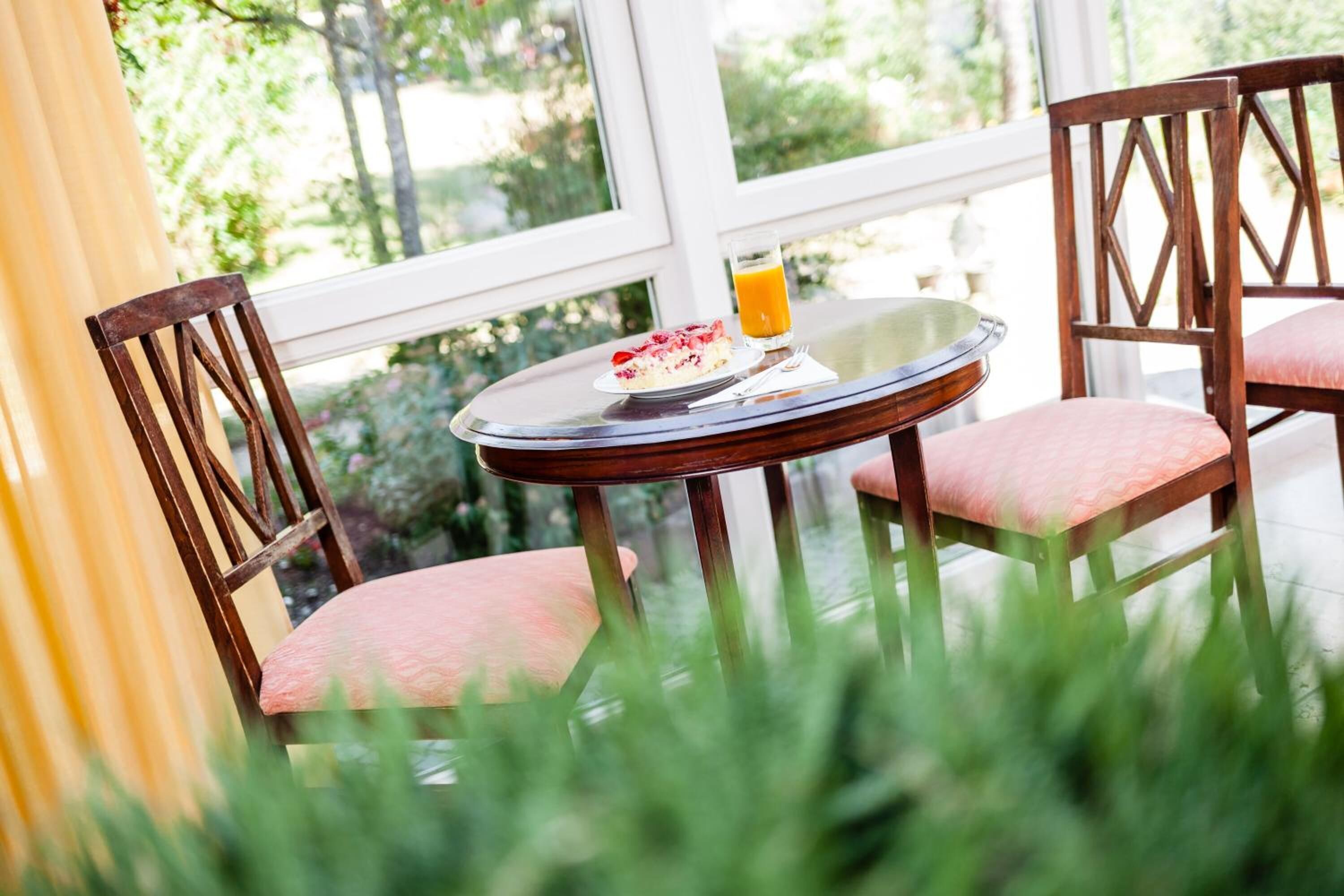 A round table with a glass of orange juice and a plate of food, surrounded by two wooden chairs with pink cushions, in a room with a large window.