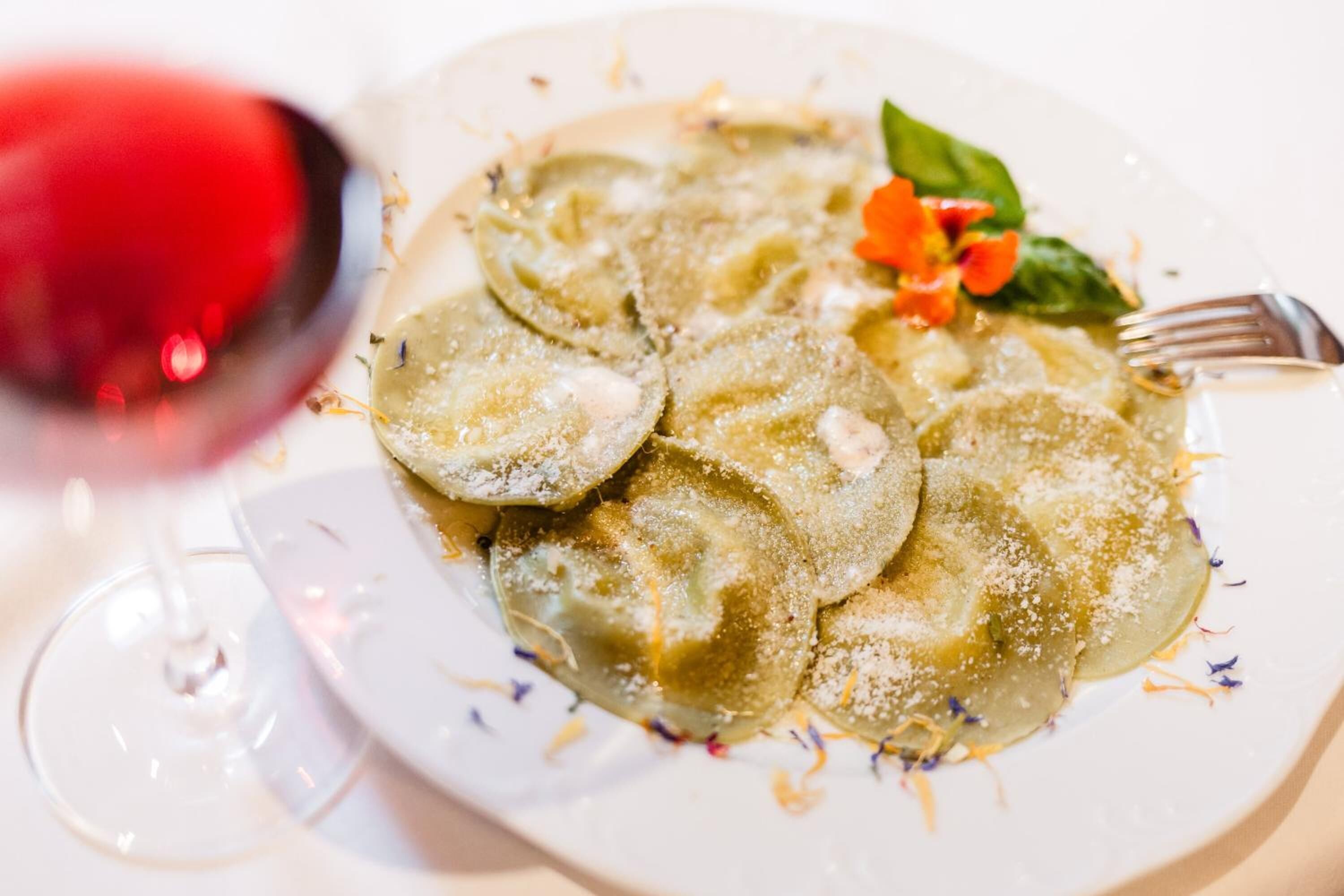 A plate of ravioli with a garnish of herbs and a small flower, accompanied by a glass of red wine.