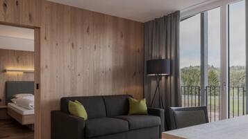A modern hotel room with a wooden wall, a dark sofa with green cushions, a bedside table, a lamp, and a balcony with a view.