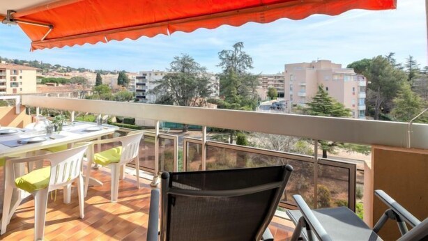 A balcony with a red awning, white chairs, a table, and a view of surrounding buildings. - Le Maxime by Interhome (Sainte Maxime)