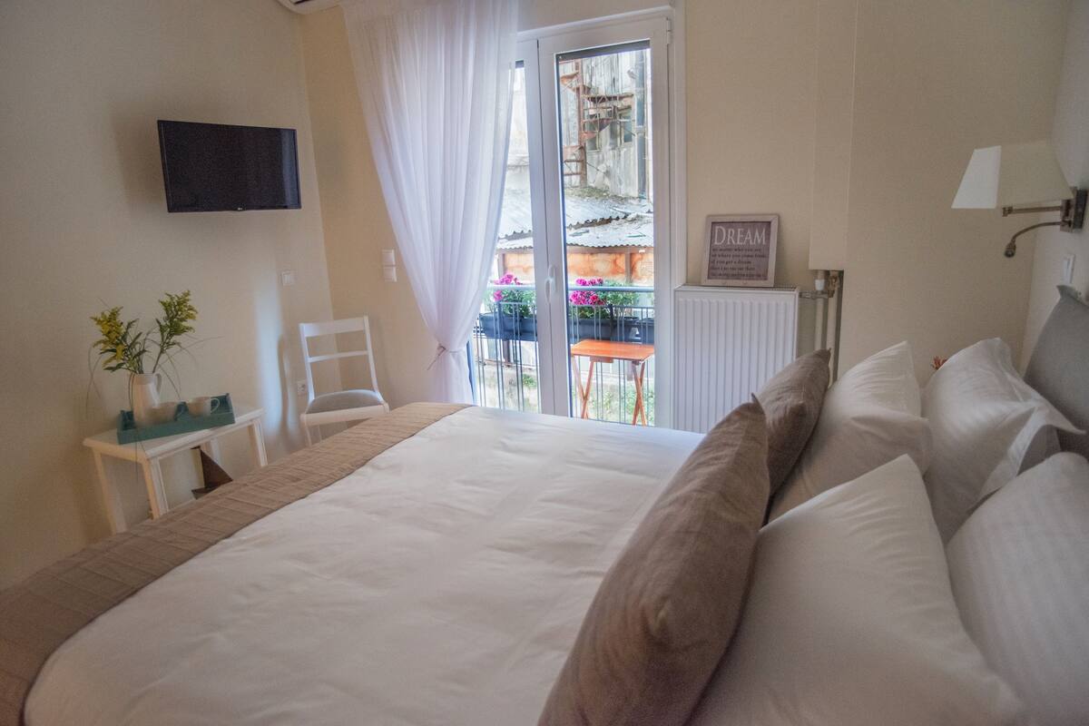A bedroom with a large bed, a television mounted on the wall, a chair, a small table with a plant, and a view of the outside balcony.