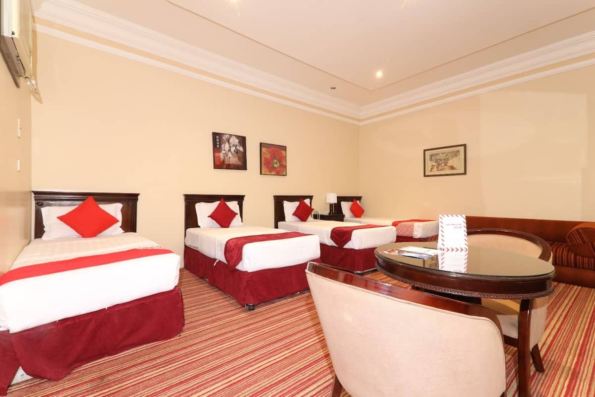 A hotel room with three beds, each with red bedding and pillows, a round wooden table, and a single armchair.