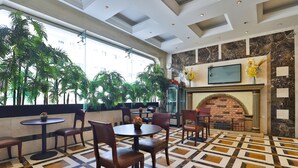 Un hall d’hôtel doté d’un sol à motifs, de différents espaces aménagés pour s’asseoir, d’une cheminée et de grandes fenêtres ornées de plantes.