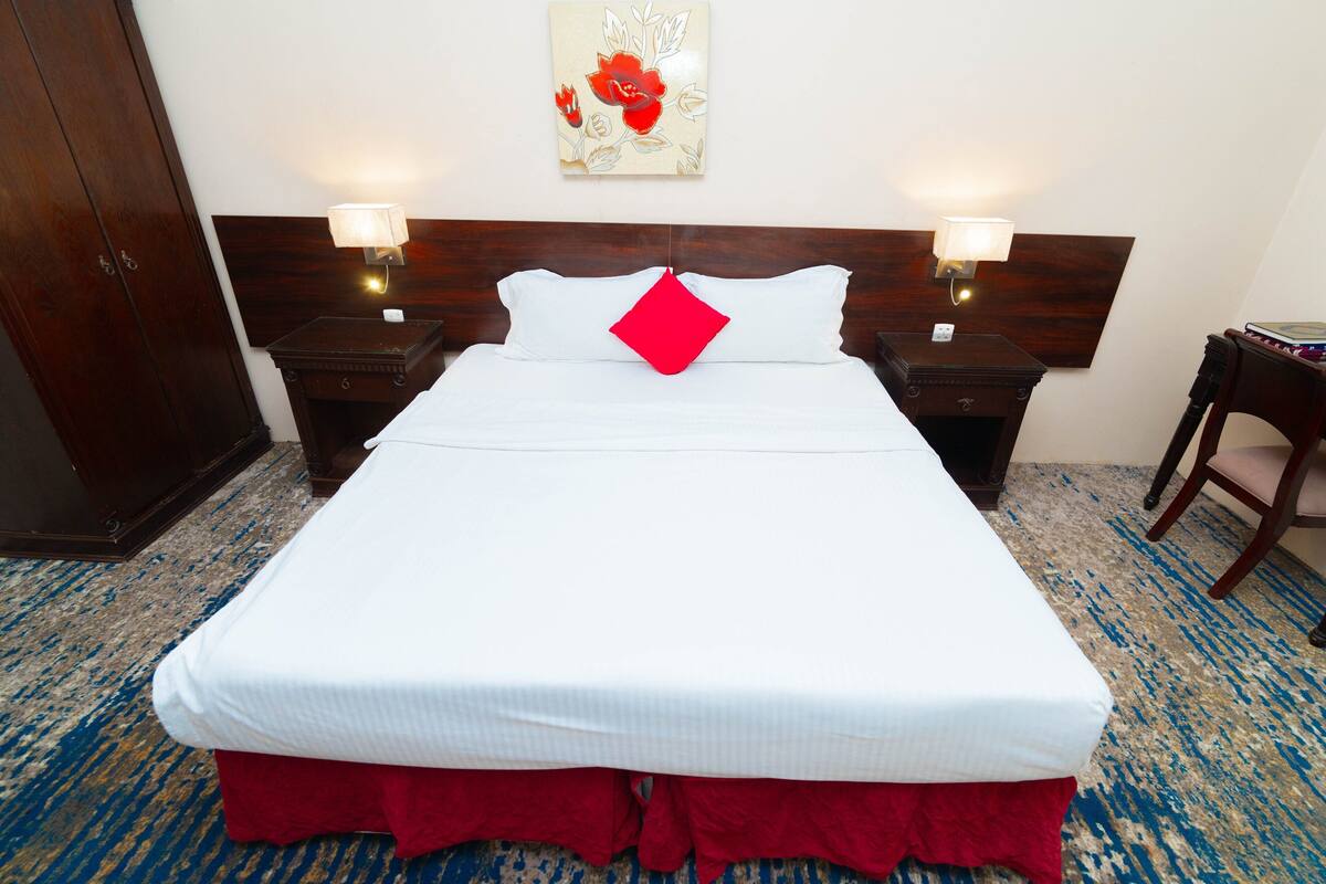 A hotel room with a large bed, two bedside tables with lamps, a wooden headboard, a red accent pillow, a floral artwork on the wall, and a blue patterned carpet.