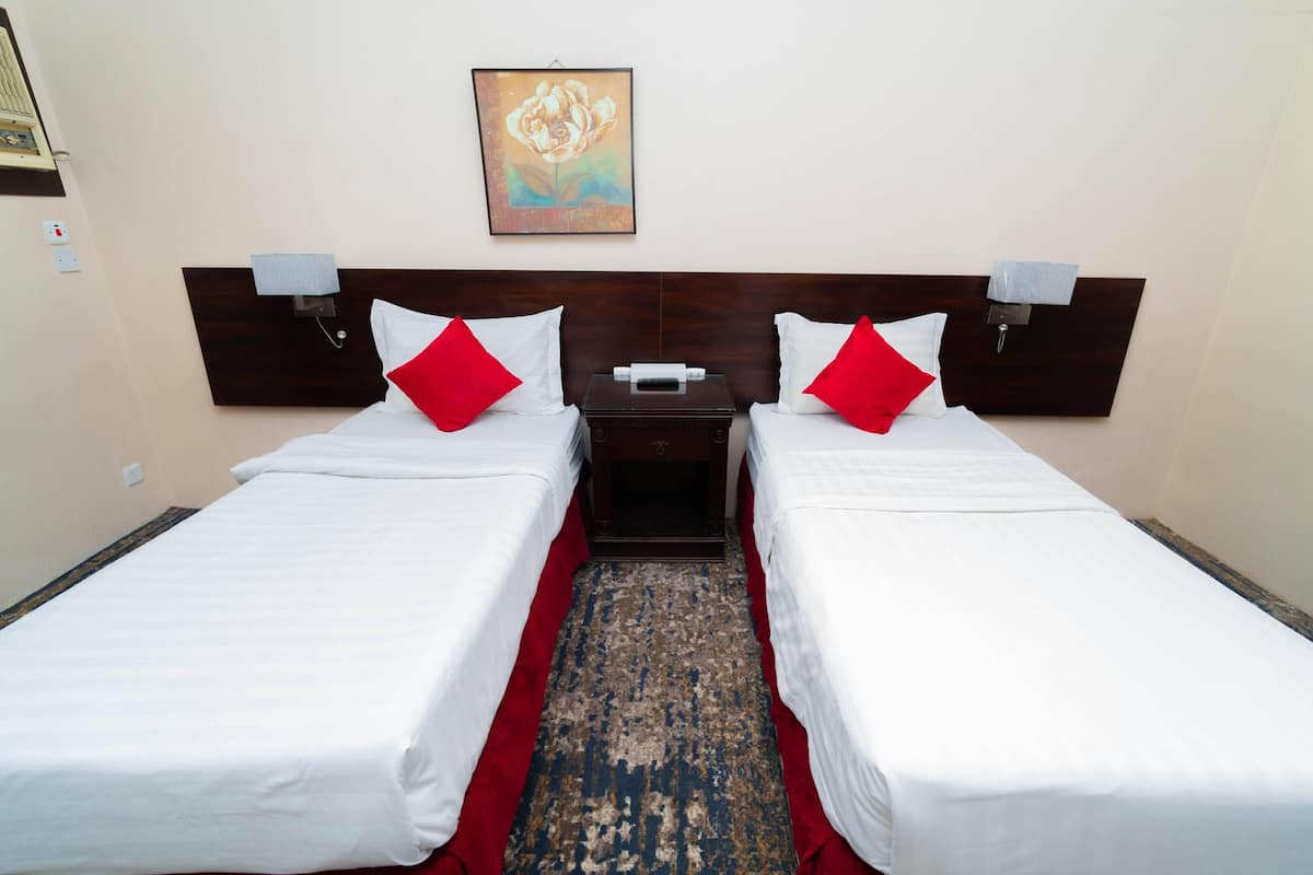 A hotel room with two single beds, each with white bedding and red pillows, a wooden headboard, a small bedside table with a lamp, and a painting on the wall.