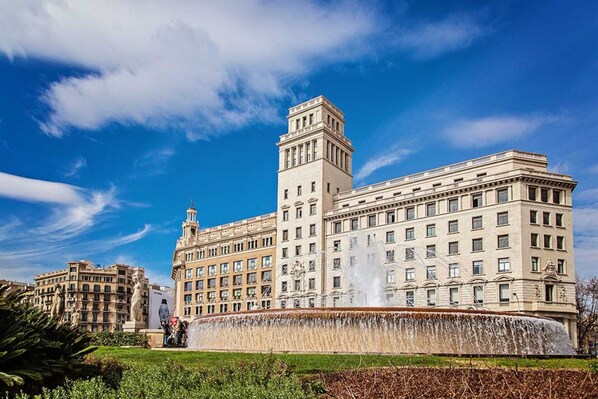 A large, historic building with a central tower and multiple windows, surrounded by a fountain and greenery.