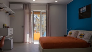 A bedroom with a bed, a desk, and a chair. There is a window with curtains, a wall-mounted air conditioner, and a view of buildings outside. - Montedonzelli B&B (Naples)