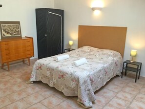 En snyggt bäddad säng med ett blommigt överkast och två hoprullade handdukar, en byrå i trä, en svart garderob och två sänglampor.