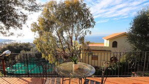 Eine Terrasse mit Glastisch und Stühlen, ein Schwimmbad und Blick auf das Meer sowie ein Gebäude mit rotem Dach.