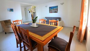 Ein Essbereich mit einem rechteckigen Tisch, sechs Holzstühlen, einer Blumenvase und einer Couch im Hintergrund.