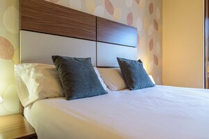 A neatly made bed with white and grey bedding, a wooden headboard, and a wall with a leaf pattern.