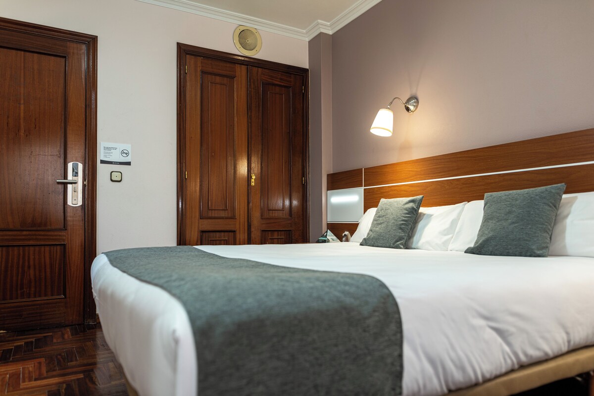 A hotel room with a large bed, wooden wardrobe, and a wall-mounted light fixture.