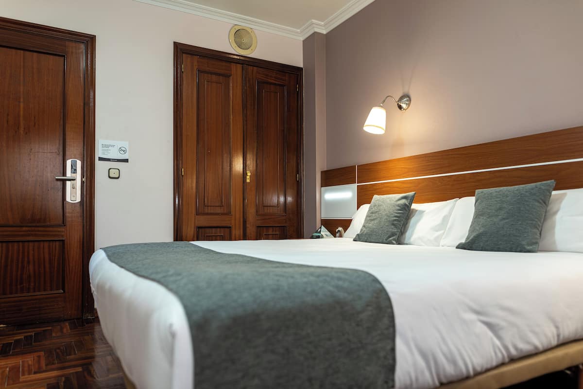 A hotel room with a large bed, wooden wardrobe, and a wall-mounted light fixture.