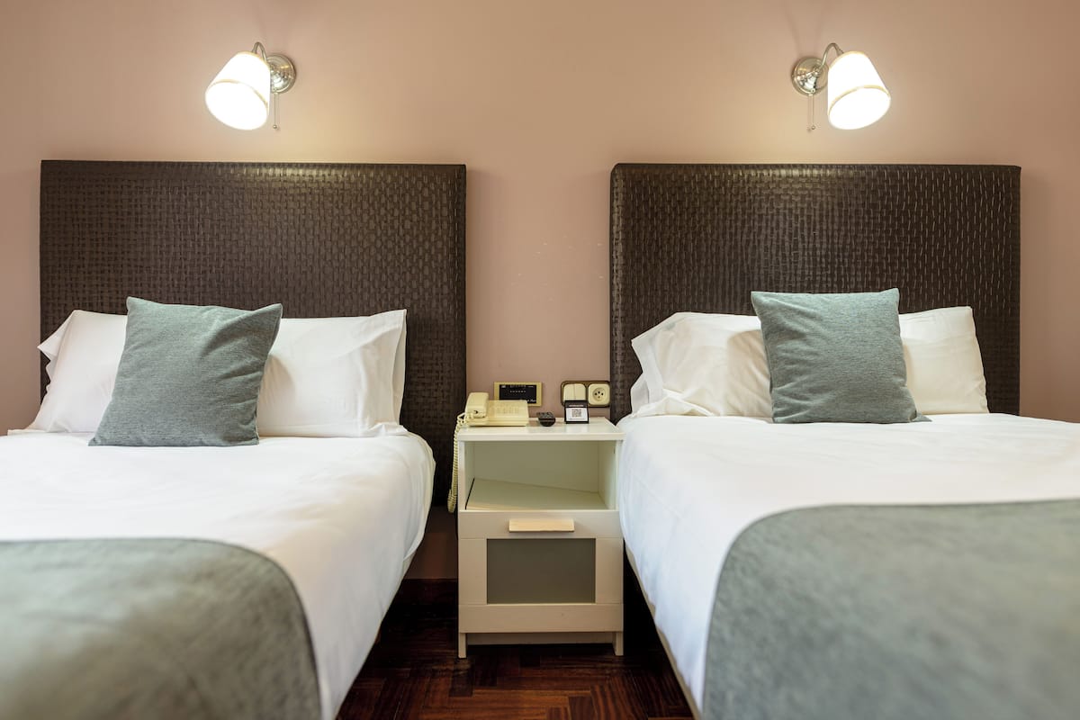 A hotel room with two beds, each with white bedding and a grey pillow. There are bedside tables with lamps and a telephone.