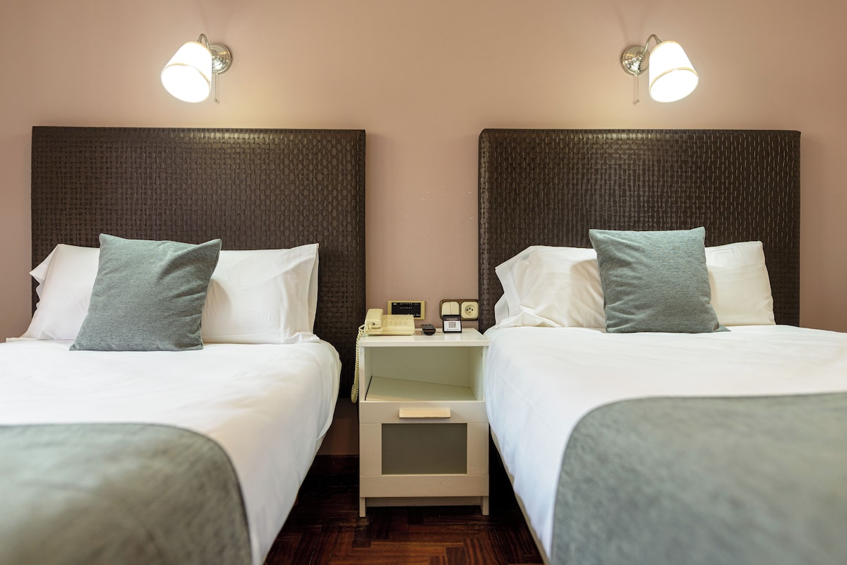 A hotel room with two beds, each with white bedding and a grey pillow. There are bedside tables with lamps and a telephone.