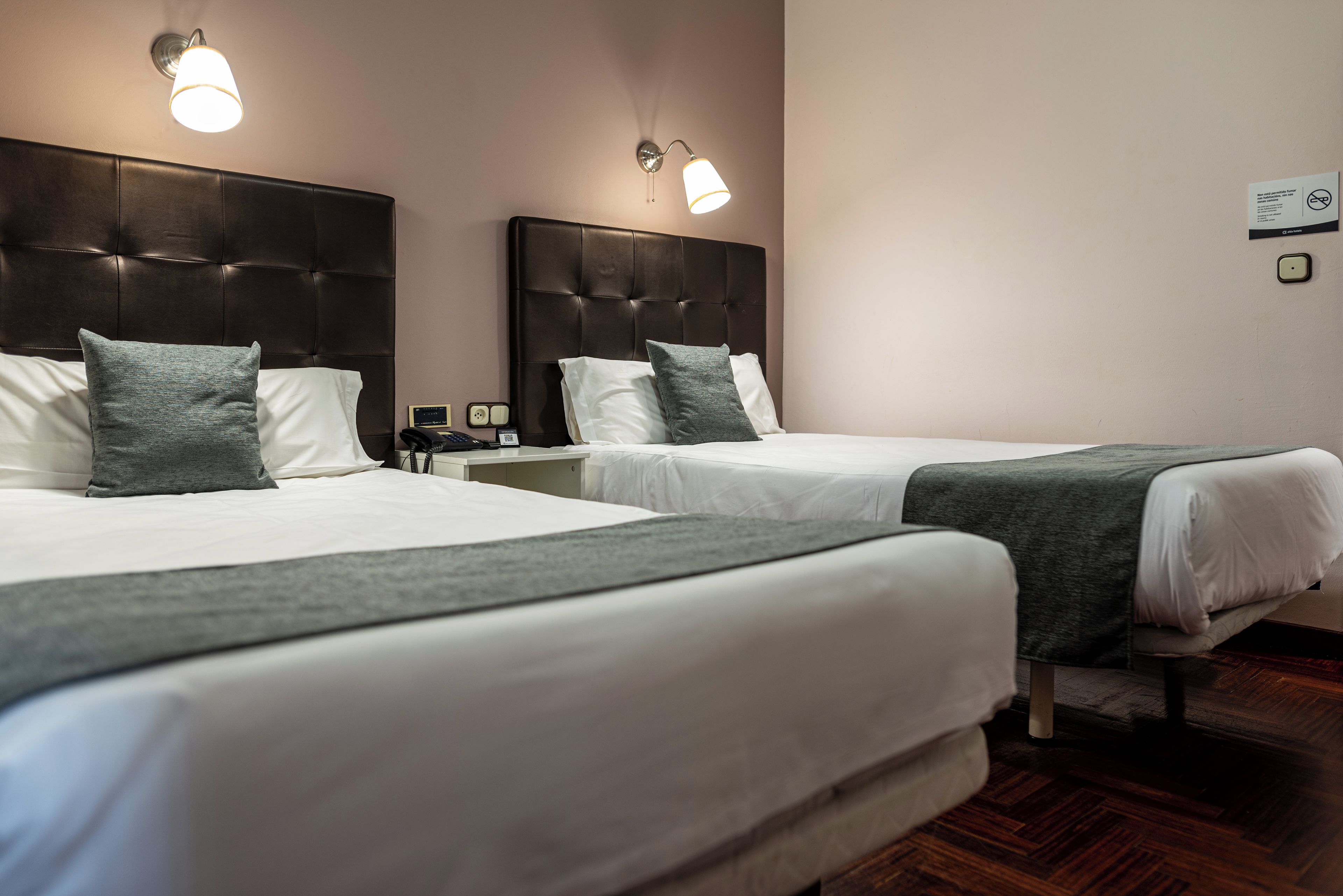 A hotel room with two beds, each with a headboard, white linens, and a grey throw blanket. There are wall-mounted light fixtures above each bed.