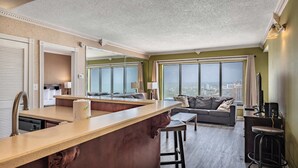 A modern hotel room with a kitchen area, a dining table, a sofa, and large windows offering a city view. - Coastal Luxe | Palace Twenty Oh Eight (Myrtle Beach)