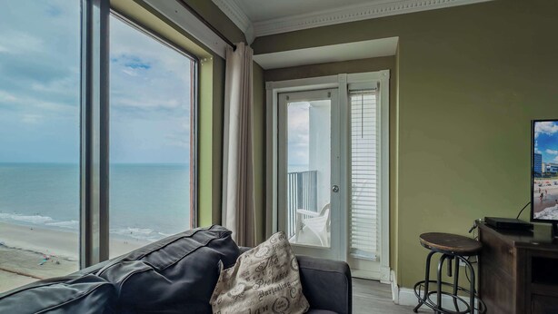 A living room with a view of the beach, a leather sofa, a television, and a bar area. - Coastal Luxe | Palace Twenty Oh Eight (Myrtle Beach)
