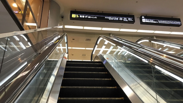 An indoor escalator with glass sides and overhead signs indicating directions to various terminals. - First Cabin Haneda Terminal 1 (Tokyo)