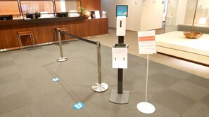 Un área de recepción con mostrador de madera, una pantalla digital, un letrero de distanciamiento social y un cartel que indica 'Primera cabina'.
