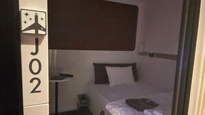 A single-bed room with a bunk bed above, a small table, a bedside lamp, and a suitcase on the bed. - First Cabin Haneda Terminal 1 (Tokyo)