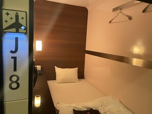 Una habitación pequeña con una cama individual, cabecera de madera, mesita de noche con lámpara y perchero.