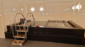 A modern bathroom with a black tiled floor, a glass-enclosed bathtub, and a metal staircase with a red object on the landing. - First Cabin Haneda Terminal 1 (Tokyo)