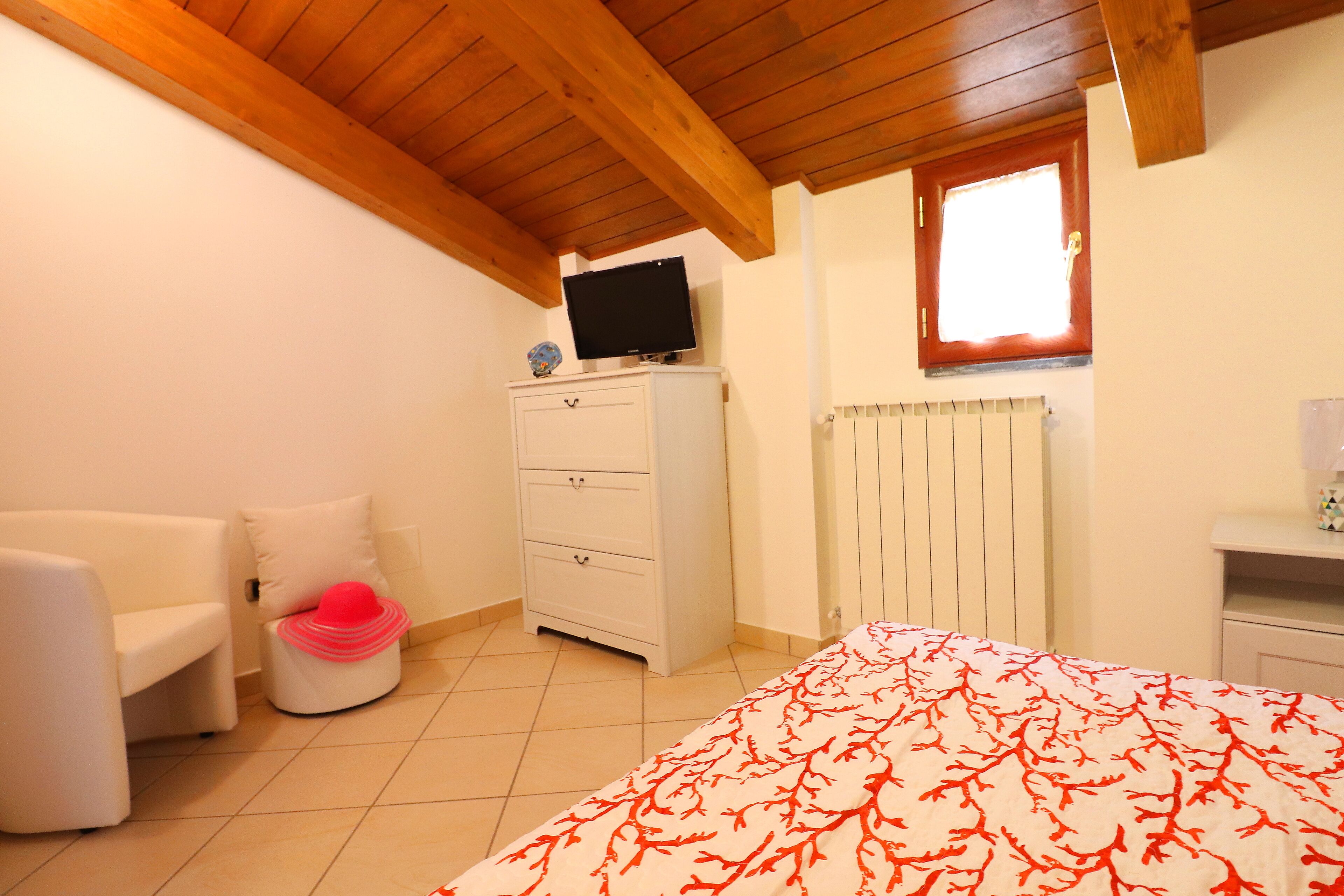 Une chambre à coucher avec un lit, une commode blanche, un téléviseur posé dessus, une fenêtre et un radiateur.