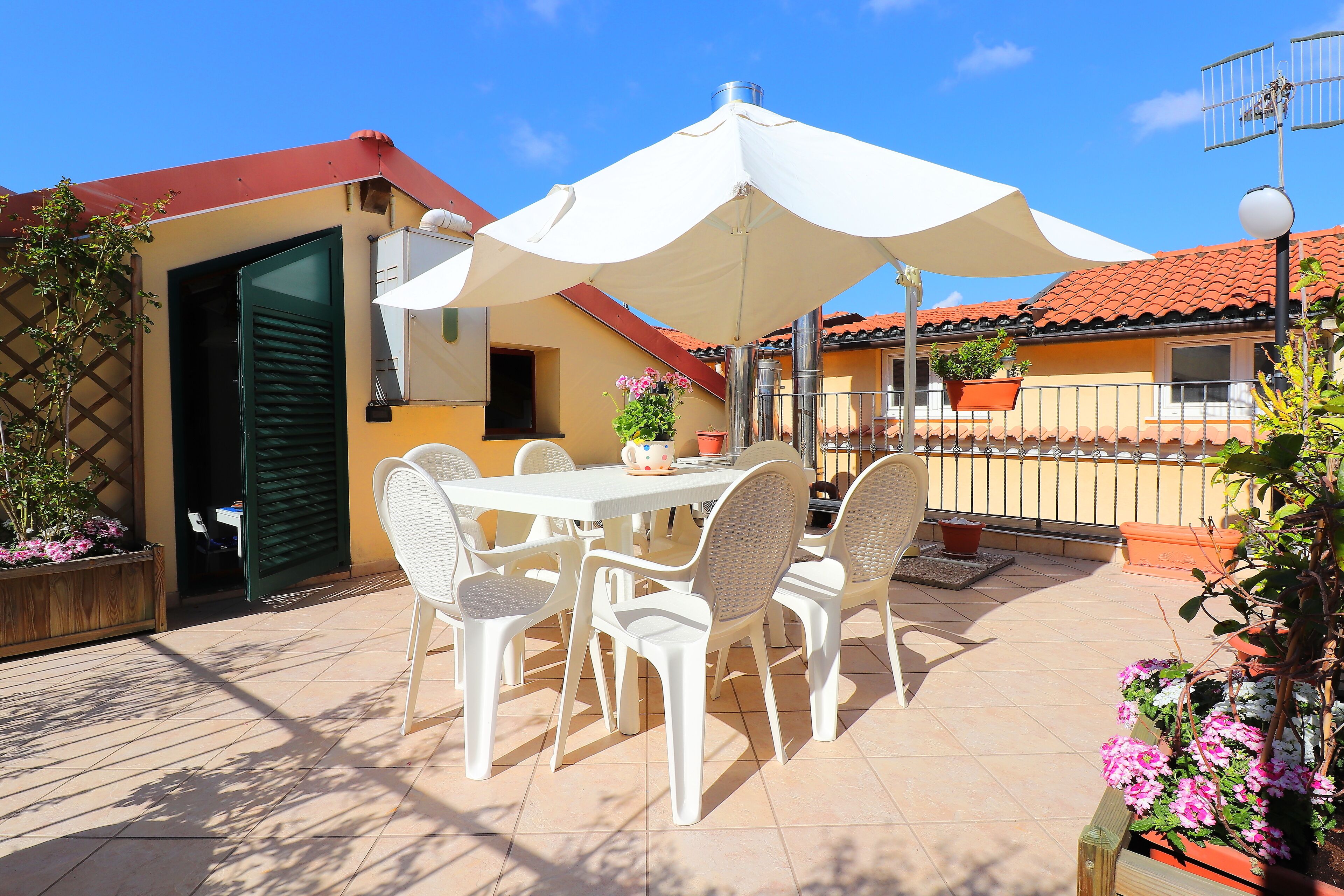 An outdoor patio with a table and chairs under a large umbrella, surrounded by potted plants and a wooden fence.