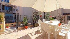 Un patio con mesa y sillas, rodeado de plantas en macetas y un paraguas grande.
