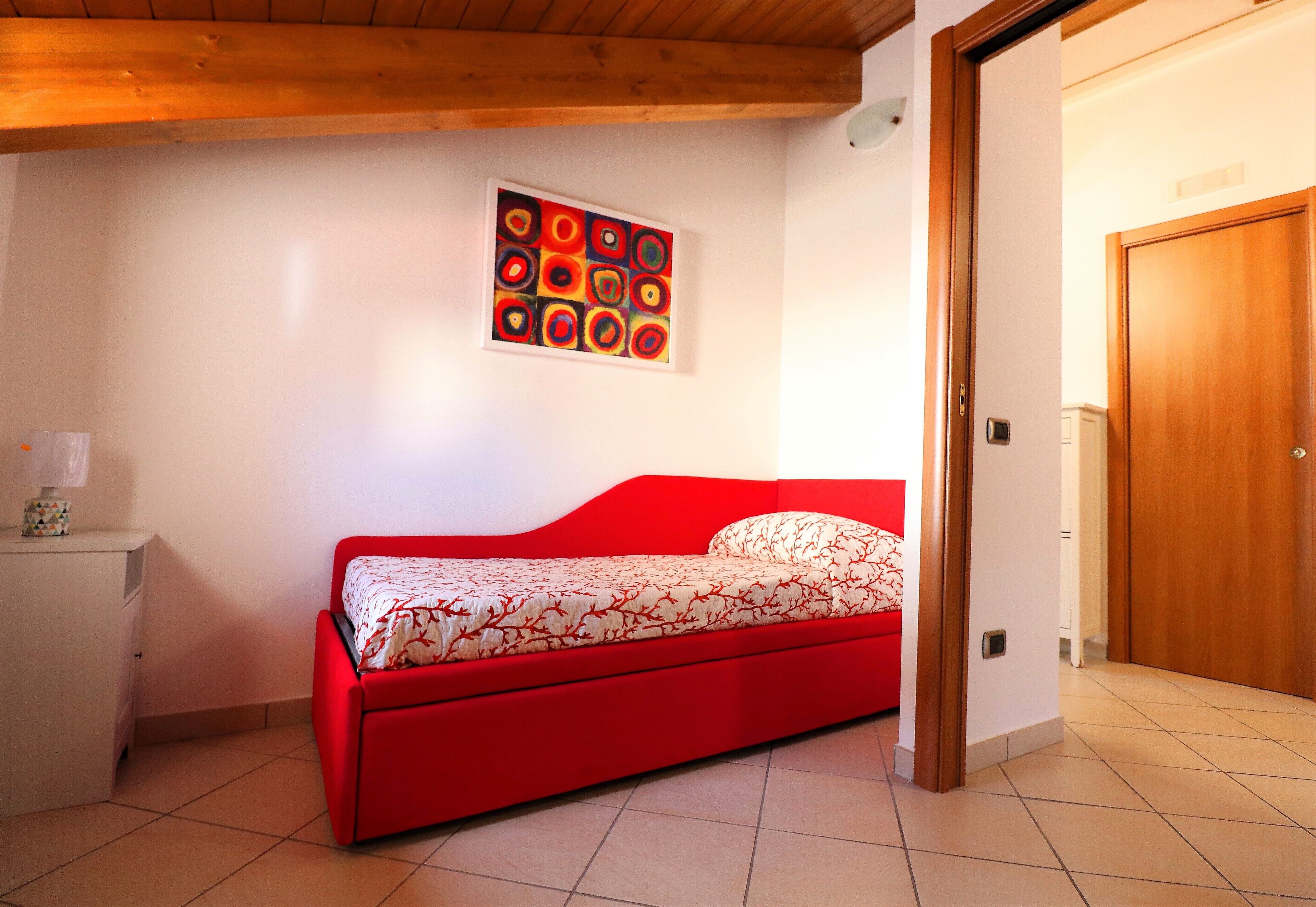 Ein kleines, rotes Bett in einem Raum mit Holzboden, einer weißen Wand und einer Tür.