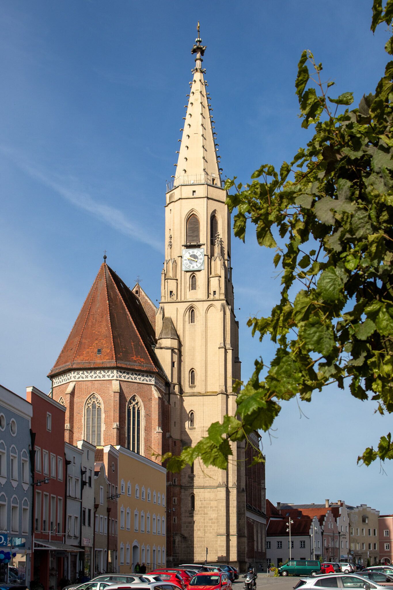 A historic church with a tall spire and a clock tower, surrounded by traditional buildings and parked cars.