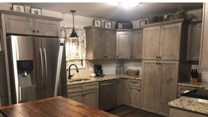 A kitchen with wooden cabinets, a stainless steel refrigerator, granite countertops, and a wooden dining table. - Greer’s Ferry Lake & Little Red River Cabin (Heber Springs)