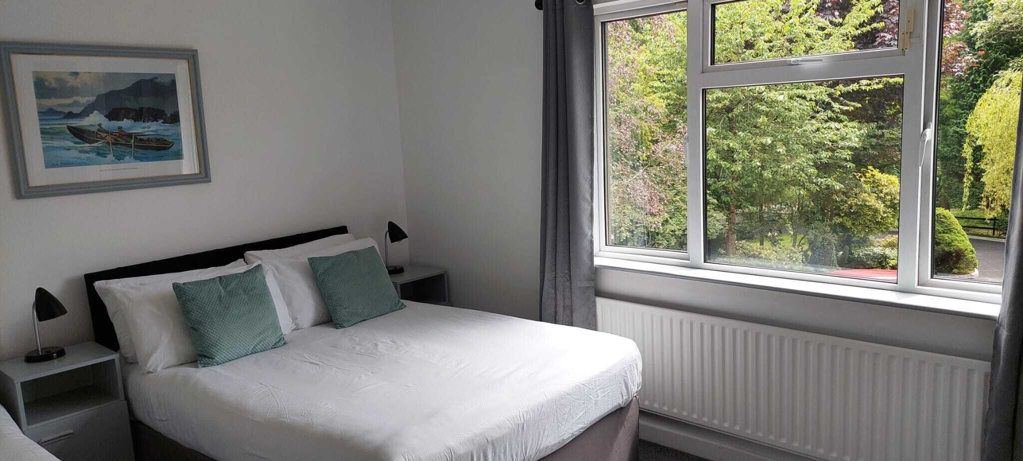 Una cama bien hecha con ropa blanca y dos almohadas verdes, una mesita de noche con una lámpara, y una ventana con vistas a vegetación y un edificio.