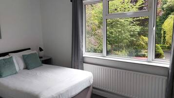 Una cama bien hecha con ropa blanca y dos almohadas verdes, una mesita de noche con una lámpara, y una ventana con vistas a vegetación y un edificio.