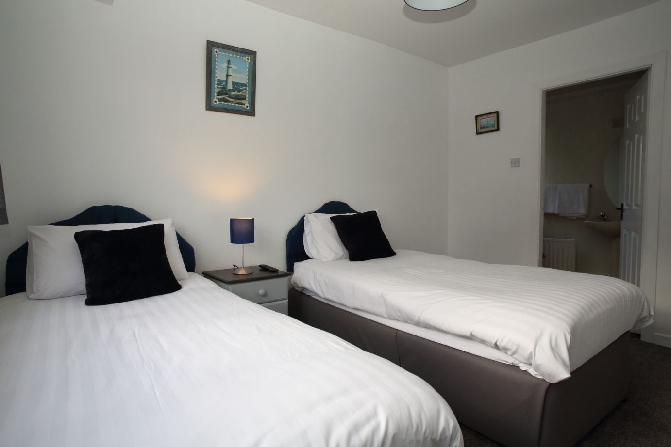 Habitación de hotel con dos camas individuales, una mesita de noche con lámpara, un cuadro enmarcado en la pared y un baño visible a través de una puerta abierta.