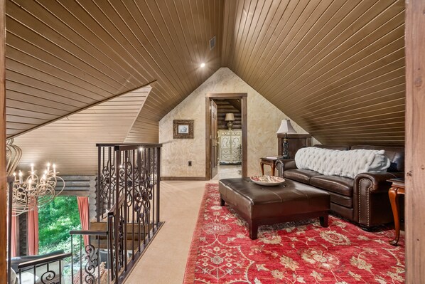 A spacious loft bedroom with a high ceiling, a large window, a cozy seating area with a leather sofa, and a red patterned rug.